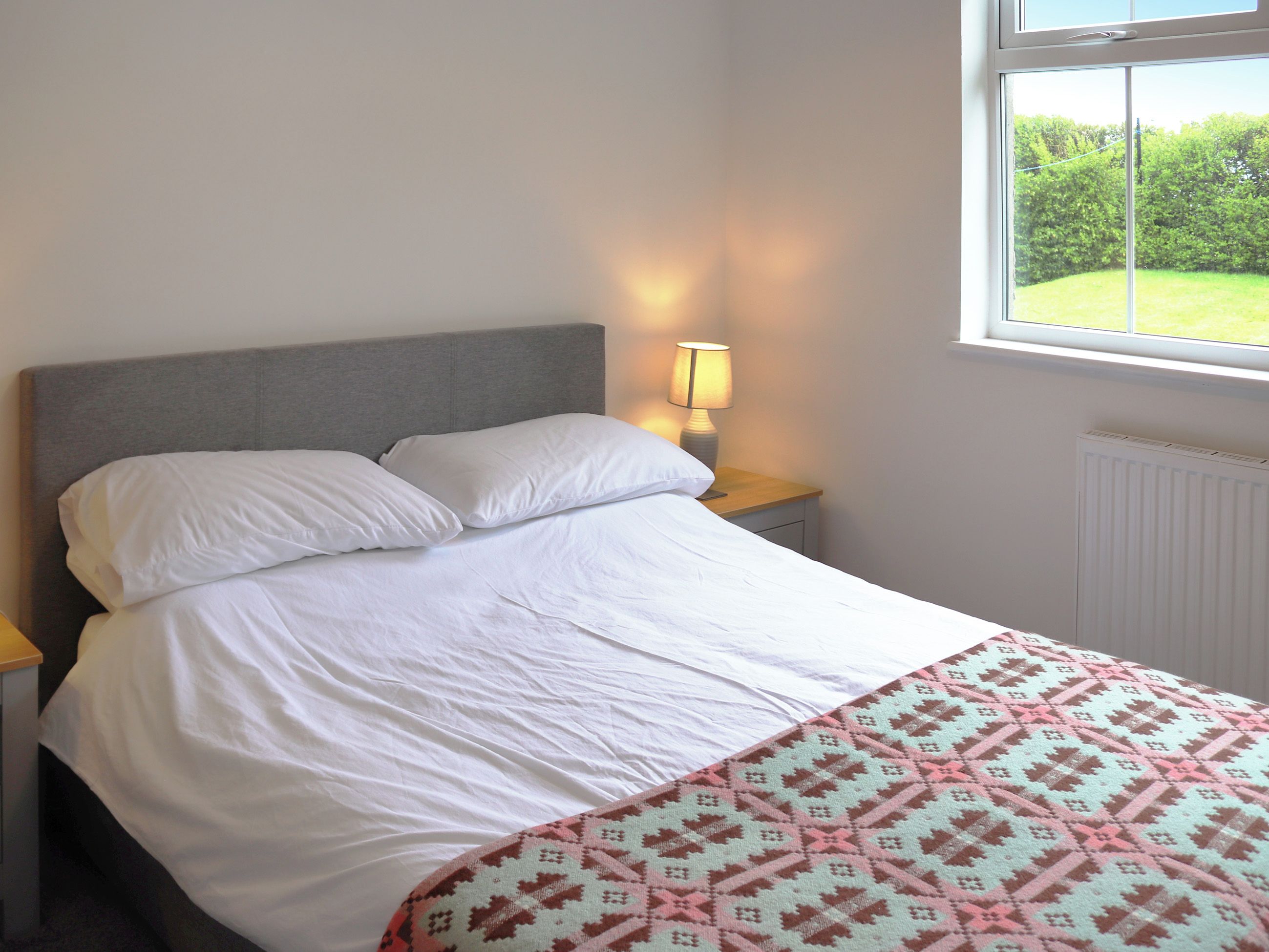 A bed with white linens, a patterned throw, a bedside lamp, and a window with a view of greenery.