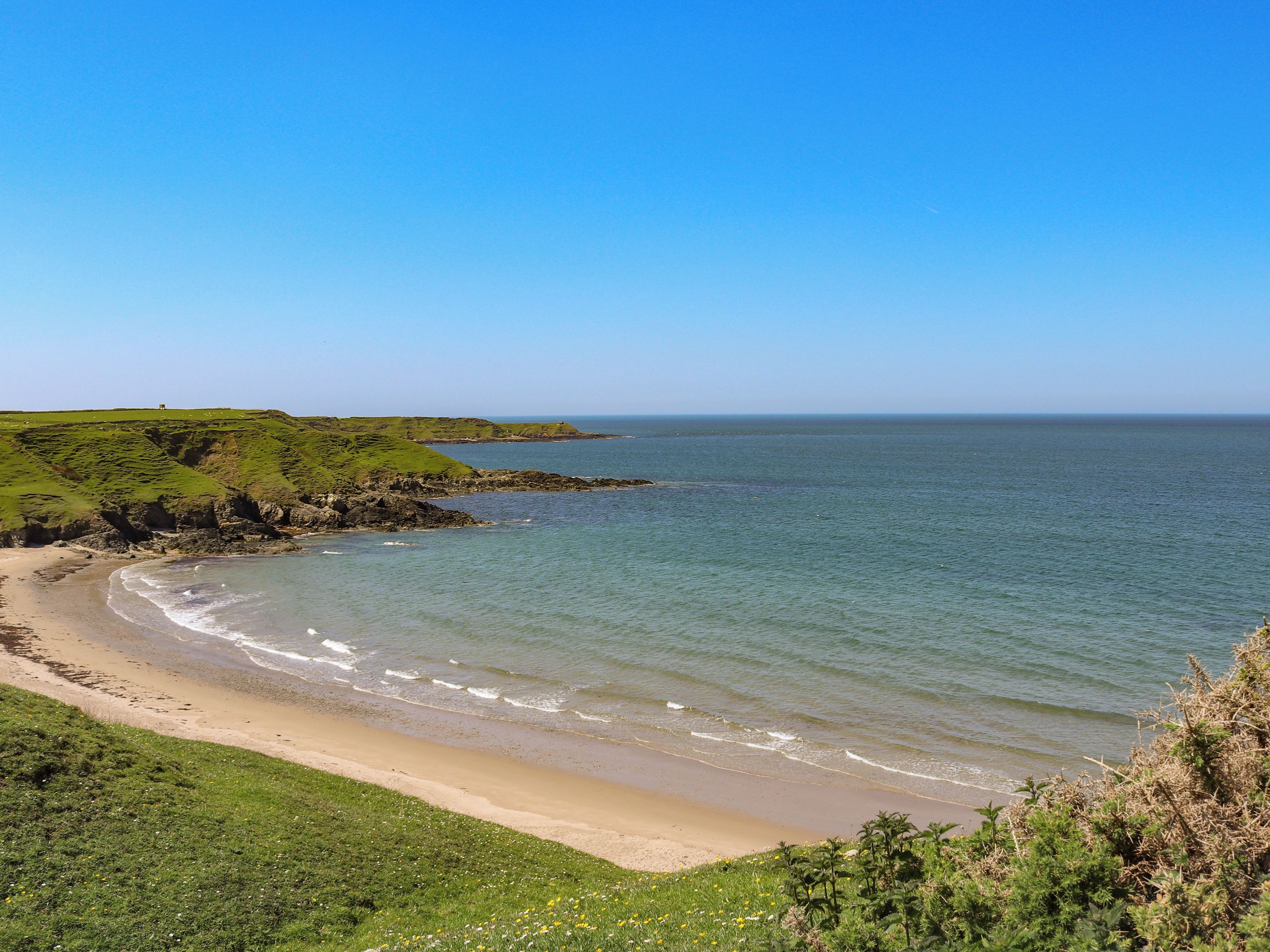 A coastal landscape with a sandy beach, grassy hills, and a clear blue sky.