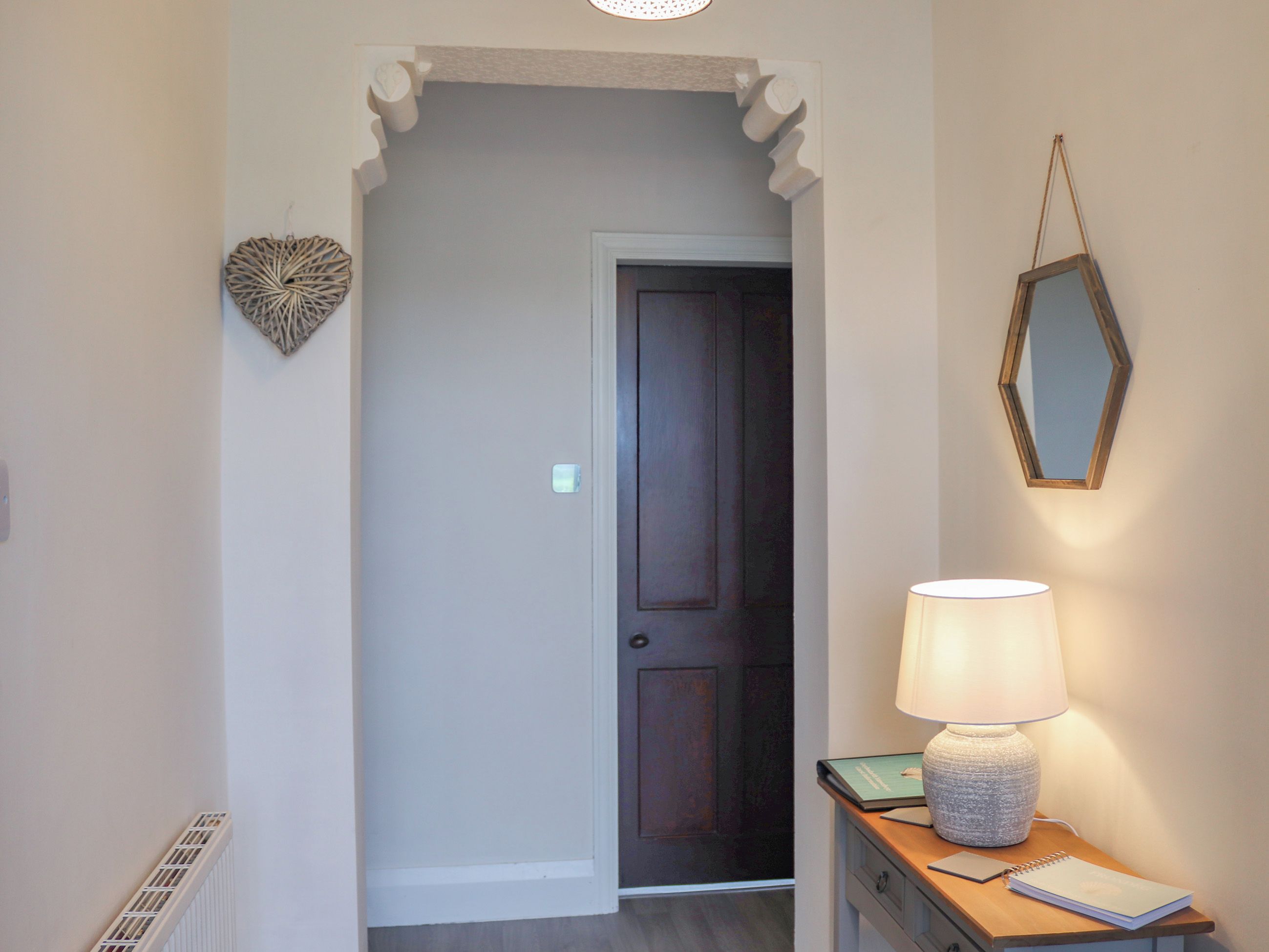 Un couloir avec une porte sombre, un décor mural en forme de cœur, un miroir et une table sur laquelle se trouvent une lampe et un carnet.