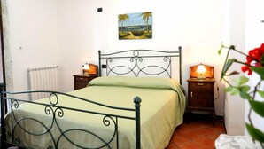 A single bed with a metal frame and a green bedspread in a room with white walls. There is a painting of a beach scene on the wall above the bed. Two bedside tables with lamps are visible. - AGRITURISMO  LA FOCAZZA (Ascea)