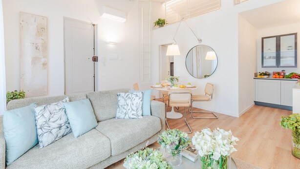 En moderne stue med en grå sofa, et spisebord med stole, et køkkenområde med skabe, og et stort spejl.