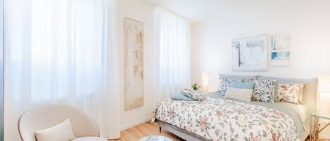 A bedroom with a bed, a chair, a small stool, and artwork on the wall.