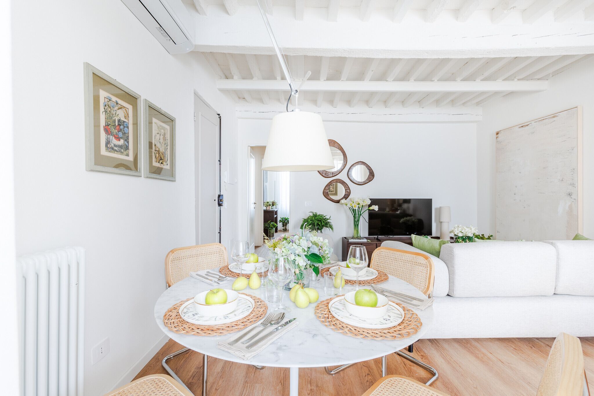 Un coin repas avec une table ronde dressée pour quatre personnes, entourée de chaises en osier, d’un canapé blanc et d’un téléviseur en arrière-plan.