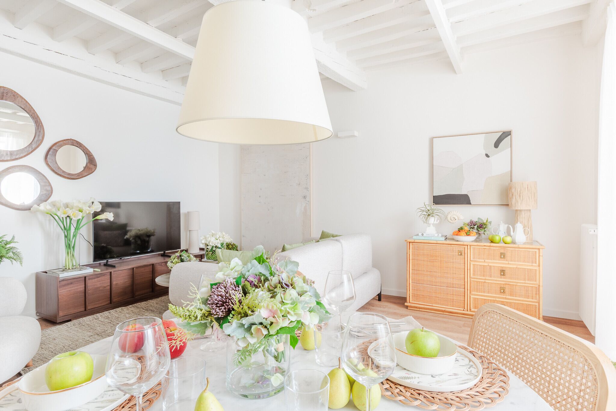 A modern living room with a white sofa, a dining table set with fruits and glasses, a TV, and a wooden cabinet.