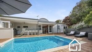 Ein Schwimmbecken mit Holzterrasse, überdachter Terrasse und Gartenmöbeln.