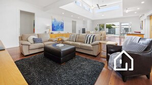 Ein geräumiges Wohnzimmer mit einer großen L-förmigen Couch, einem schwarzen Hocker und einem Ledersessel. Im Boden befinden sich ein Holztisch und ein schwarzer Teppich.