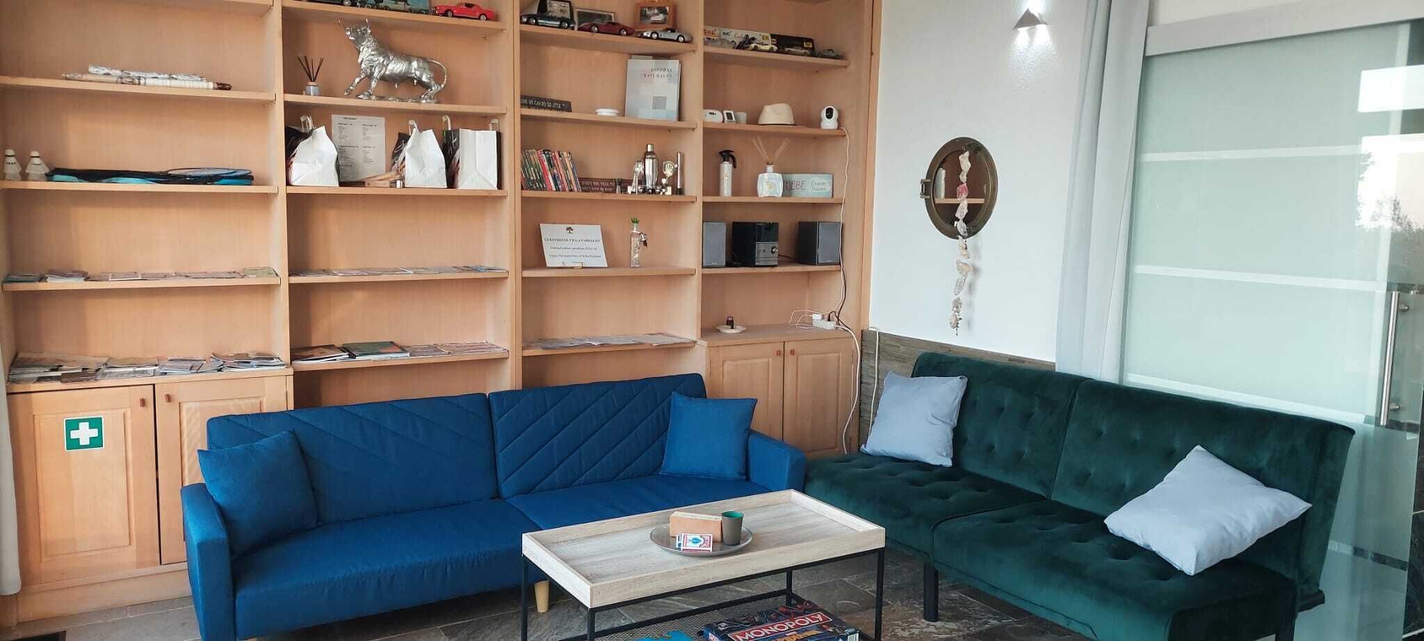 Una habitación con un sofá azul, un sofá verde, una mesa de centro y una estantería llena de objetos varios.