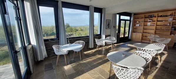 A spacious room with large windows, a dining area with white chairs and tables, and a wooden bookshelf. - Villa Pardalot Boutique Stays (Calig)