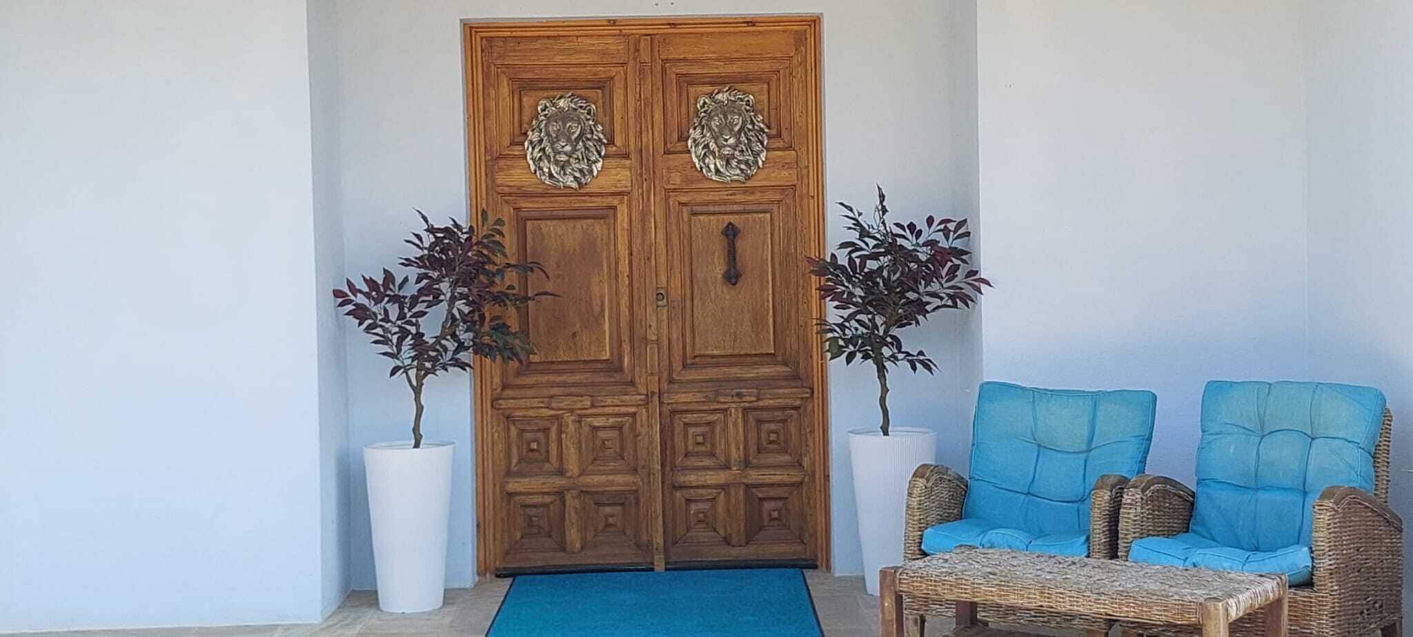 A wooden double door with lion head carvings, two potted plants, and two blue upholstered chairs with a woven table.