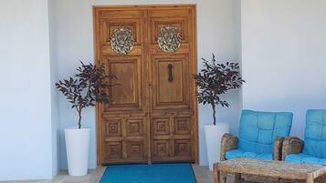 Uma porta dupla de madeira com esculturas de cabeças de leão, dois vasos de plantas e duas poltronas azuis com um mesquinho de vime.