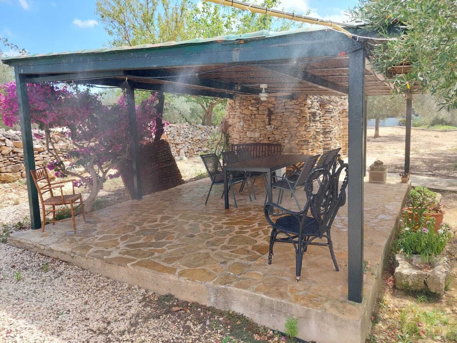 A covered outdoor patio with stone flooring, a table, chairs, and a stone wall in the background.