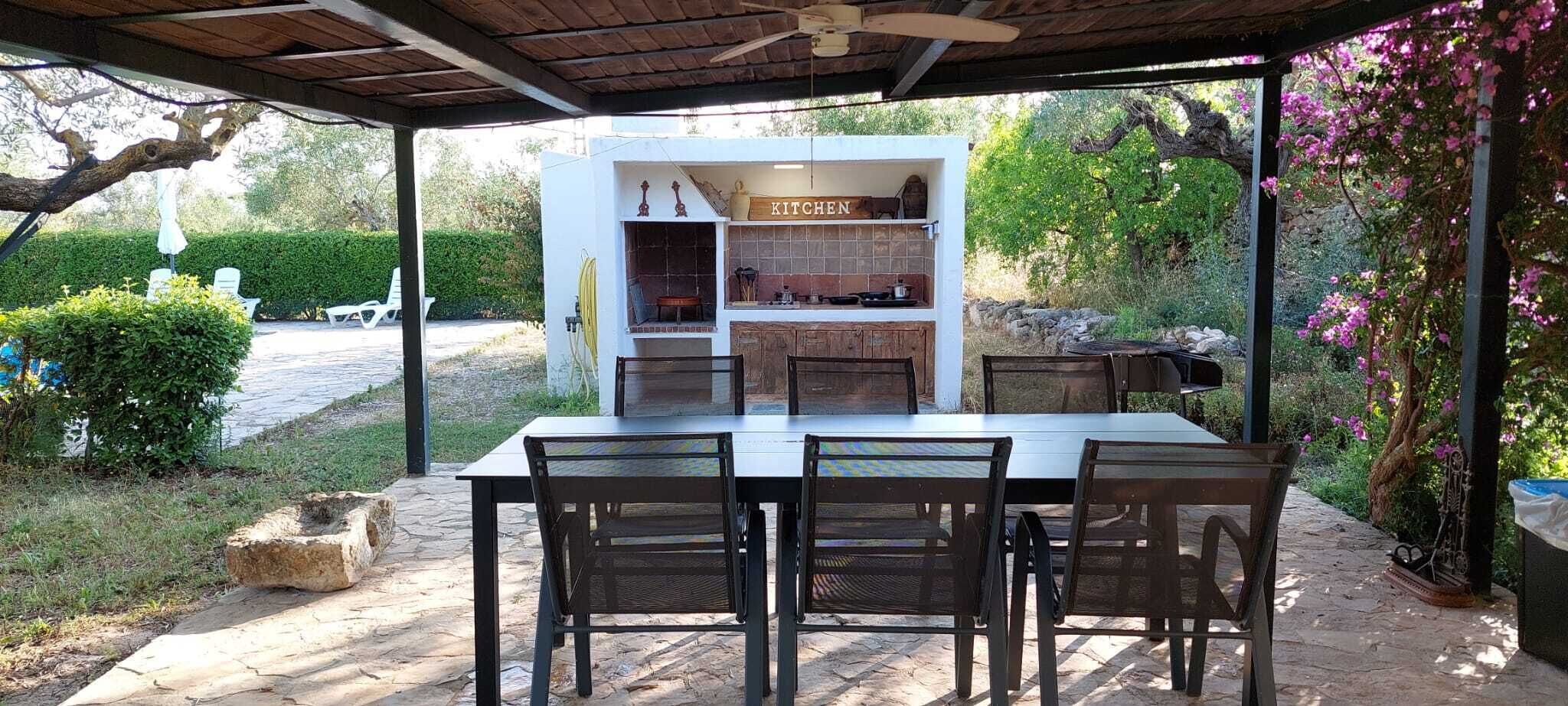 Un área de cocina al aire libre con mesa y sillas, rodeada de follaje y un sendero de piedra.