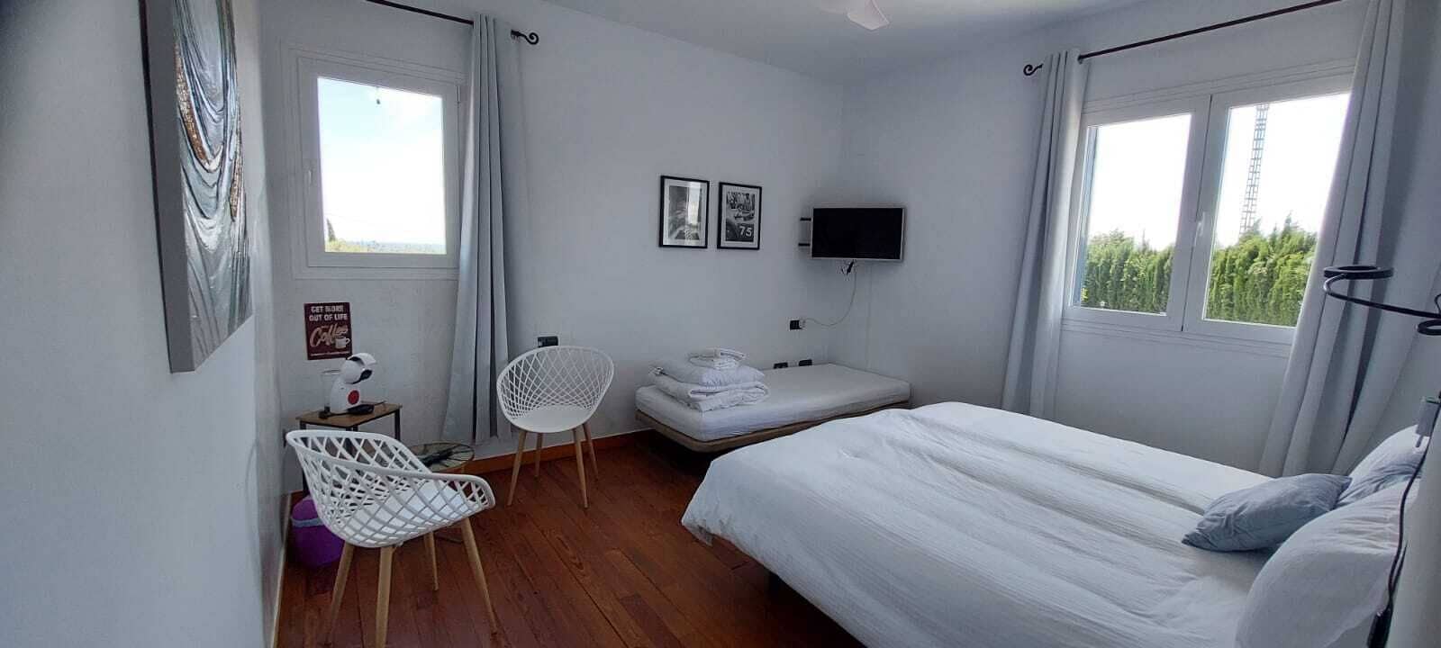 Un dormitorio con cama, una silla, una mesita, televisor y ventana.