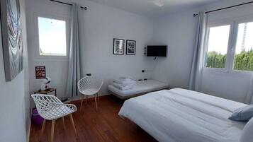 Un dormitorio con cama, una silla, una mesita, televisor y ventana.