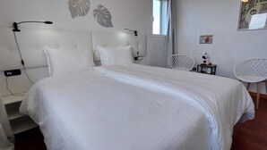 Ein ordentlich bezogenes Bett mit weißer Bettwäsche, ein Kopfteil mit einer Leuchte, ein kleiner Tisch mit einer Tasse und ein Stuhl mit Drahtgestell.