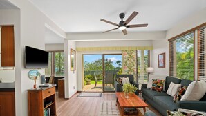 A spacious living room with a sofa, coffee table, and television. There is a sliding glass door leading to a patio with outdoor furniture. - Summer Deal Poipu Beach Condo 5 Min Walk to Ocean Pools and Hot Tub (KOLOA)