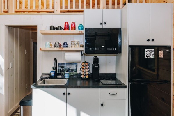 Ett kompakt kök med vita skåp, en svart mikrovågsugn, ett svart kylskåp och en kaffebryggare. Det finns hyllor med dekorativa muggar och en liten papperskorg.
