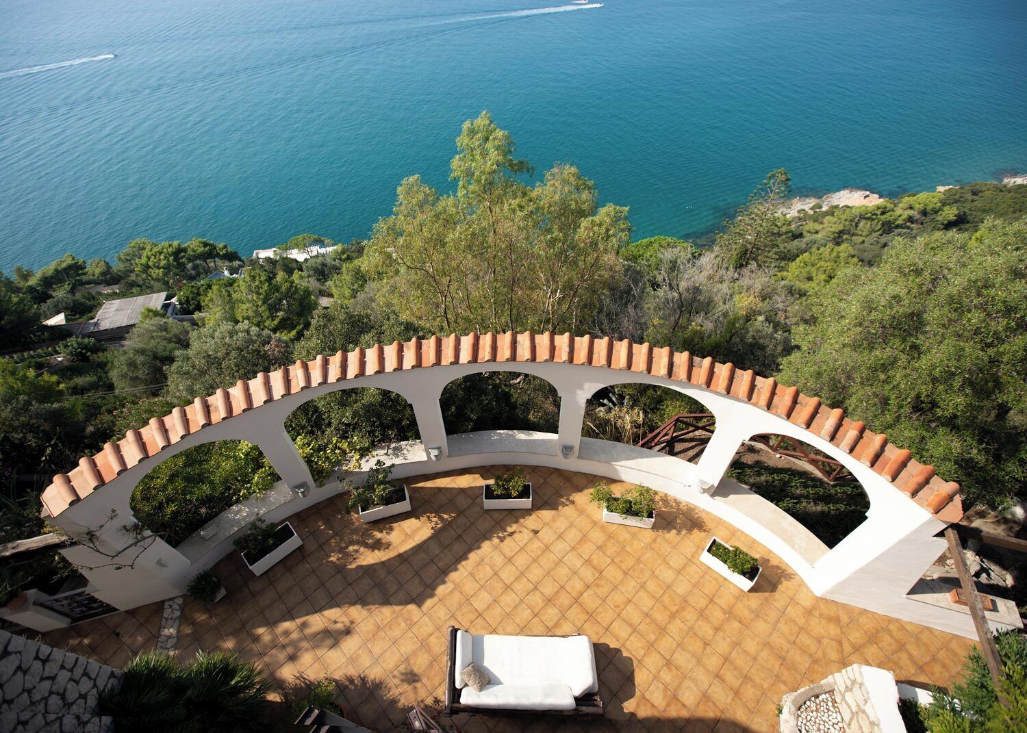A circular outdoor patio with a white tiled floor, surrounded by a white railing with terracotta tiles. There are several potted plants and a white lounge chair. The patio overlooks a body of water and a lush green landscape.
