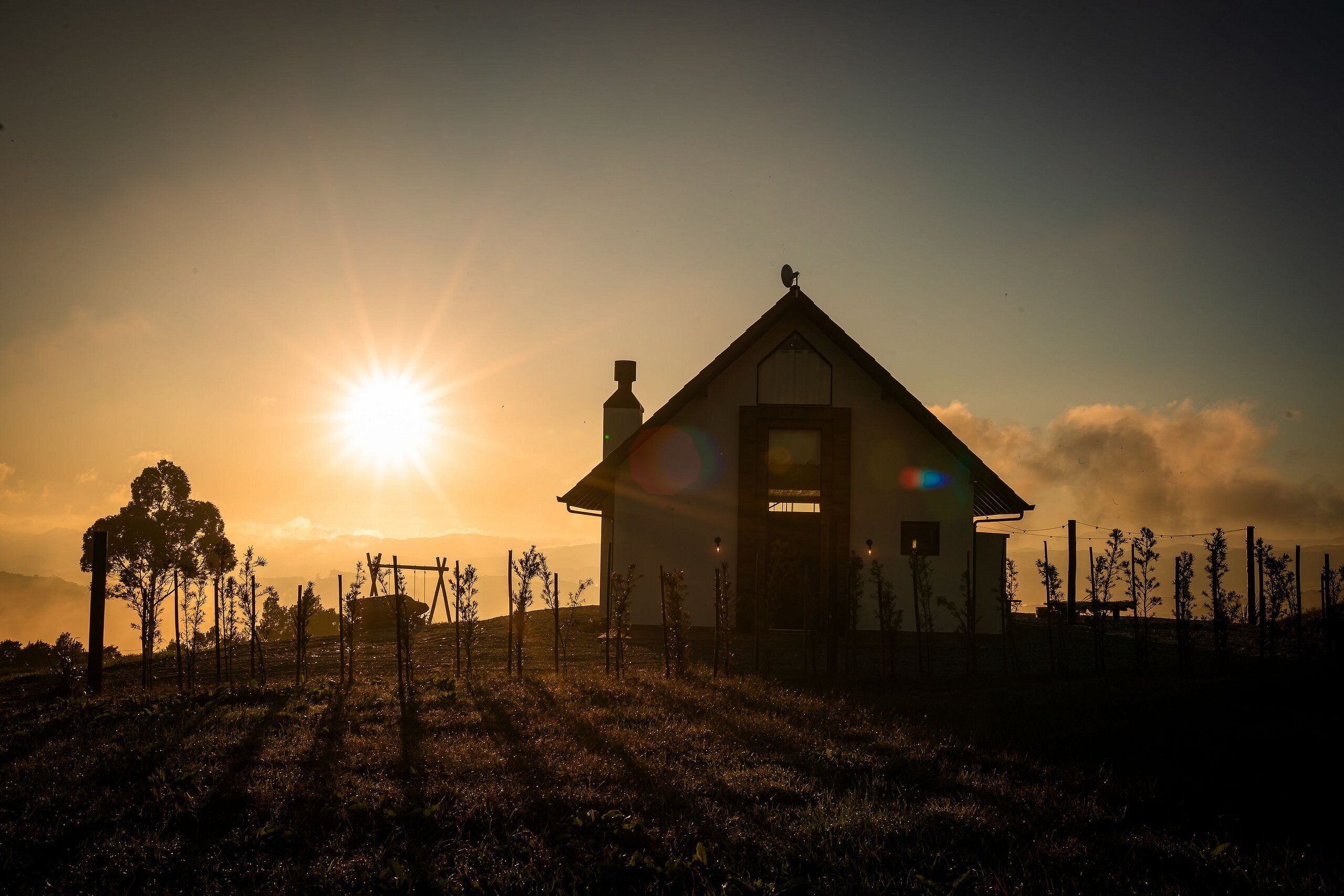 A house with a triangular roof and a chimney, surrounded by a fence and trees, with the sun setting in the background.