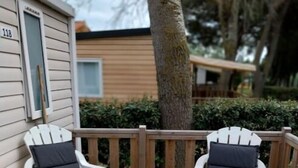 Two white Adirondack chairs with dark cushions on a wooden deck, adjacent to a beige house with a visible door number 118. - Mobilhome. (Torreilles)