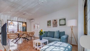 Un salon moderne avec un canapé, un coin repas avec des chaises en osier, une petite table surmontée d’un vase et des œuvres d’art accrochées aux murs.