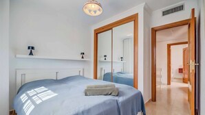 Une chambre avec un grand lit, une table de chevet avec une lampe, une armoire avec un miroir et un couloir menant à une autre pièce.