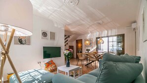 Un salon moderne avec un canapé, un téléviseur, une petite table ornée d’un vase, et un coin repas à l’arrière-plan.