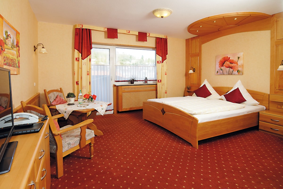 A hotel room with a wooden bed, a desk with a laptop, a chair, and a small table with flowers.