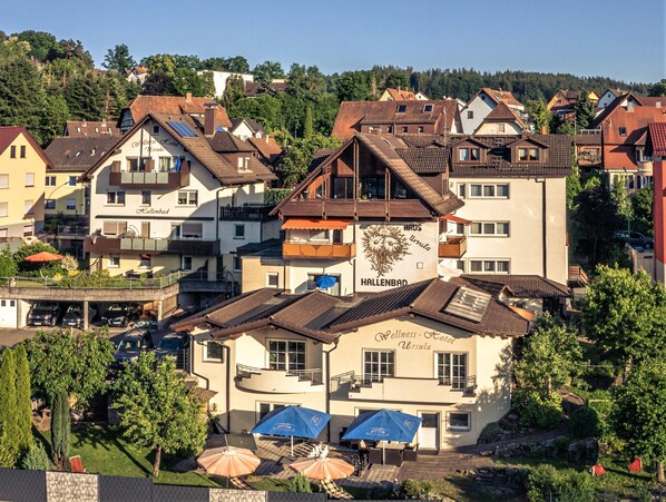 A hotel with a sign reading "HALLENBAD" and "Wellness Hotel Ursula." It features a courtyard with umbrellas and outdoor seating.