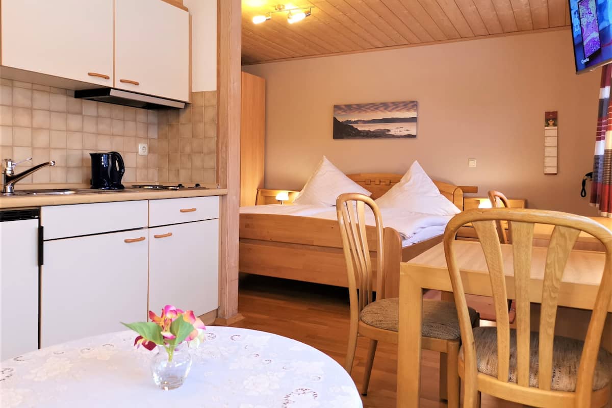 A cozy room with a wooden bed, a dining table with chairs, a kitchen area with a stove and sink, and a TV mounted on the wall.