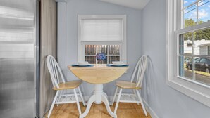 A kitchen with a stainless steel refrigerator, a wooden dining table with two chairs, a window with blinds, and a view of the outside.