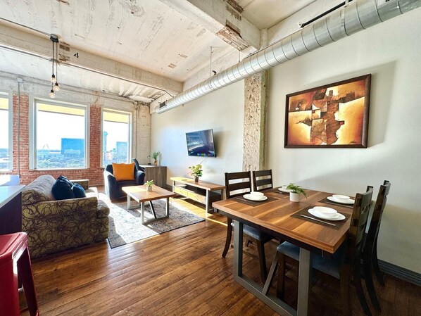 A modern living room with a dining area, large windows, a flat-screen TV, and a city view.