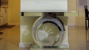 Un interior modern amb un gran mirall circular sobre un pedestal de vidre, una làmpada al sostre i una finestra amb cortines.