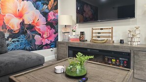 A modern living room with a large floral mural, a flat-screen TV, a fireplace, and a coffee table with a vase and remote control.