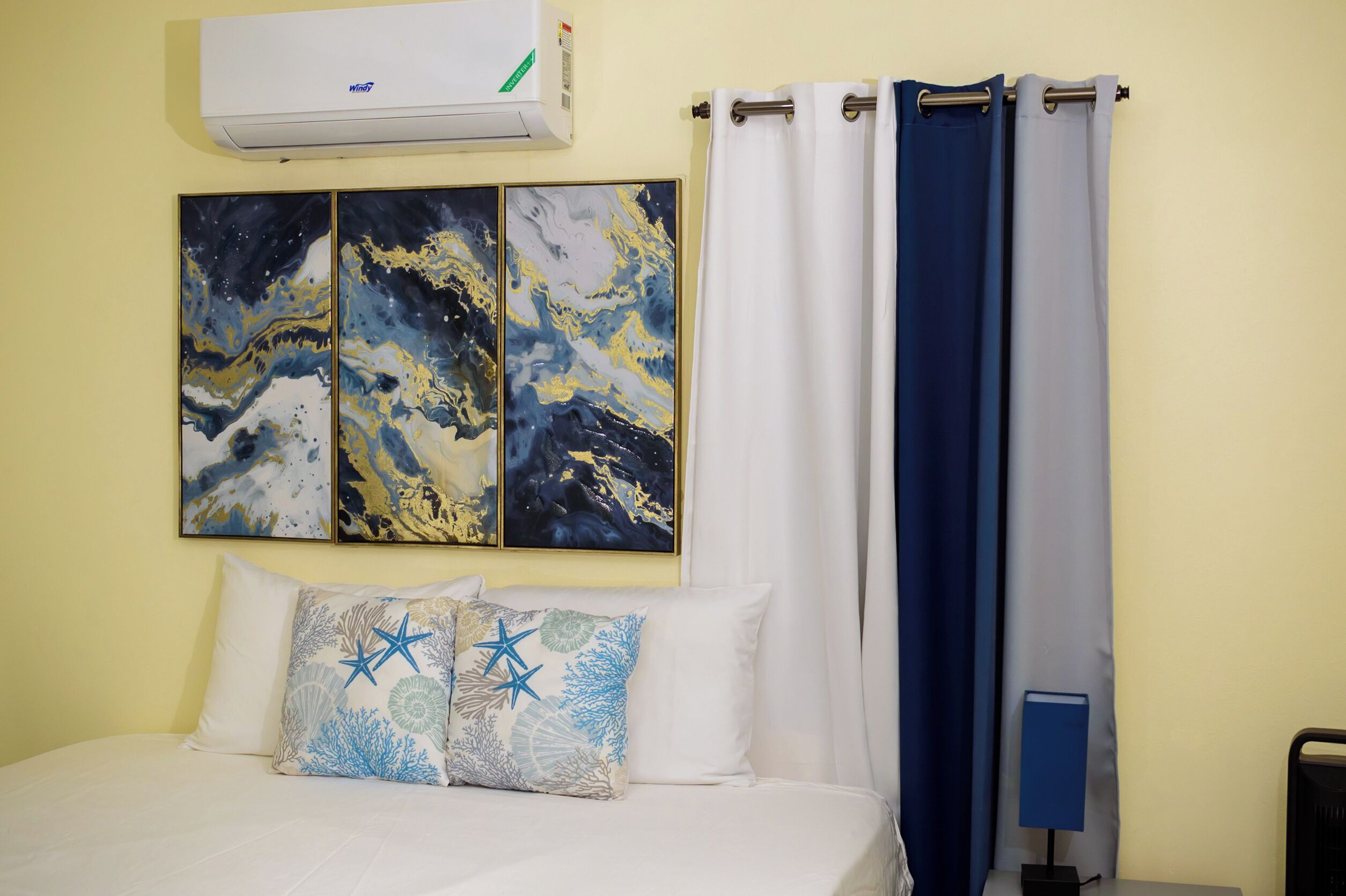 Habitación de hotel con una cama, dos almohadones decorativos, un aire acondicionado de pared y un juego de cortinas.