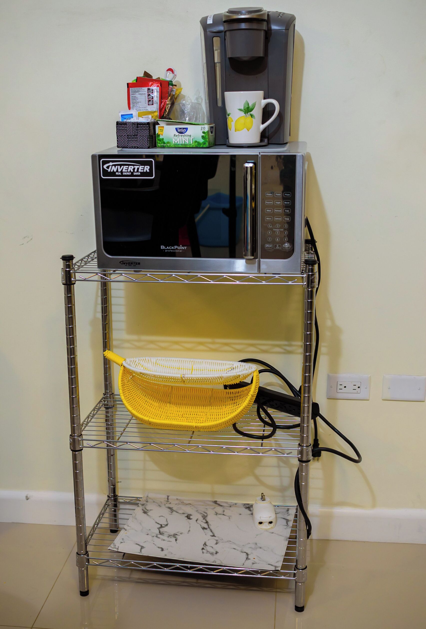 A kitchen setup with a microwave on a metal stand, a yellow basket, and a white mug on top.