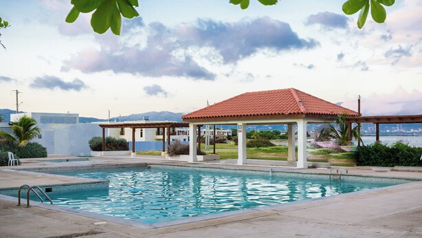 A swimming pool with a gazebo, a clear sky, and a distant view of buildings. - Beachfront Bliss in Portmore – Stylish 1BR with Ocean Views & Modern Comfort (Portmore)