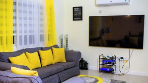 A living room with a grey sofa, yellow cushions, a flat-screen TV, and a decorative rug. - Beachfront Bliss in Portmore – Stylish 1BR with Ocean Views & Modern Comfort (Portmore)