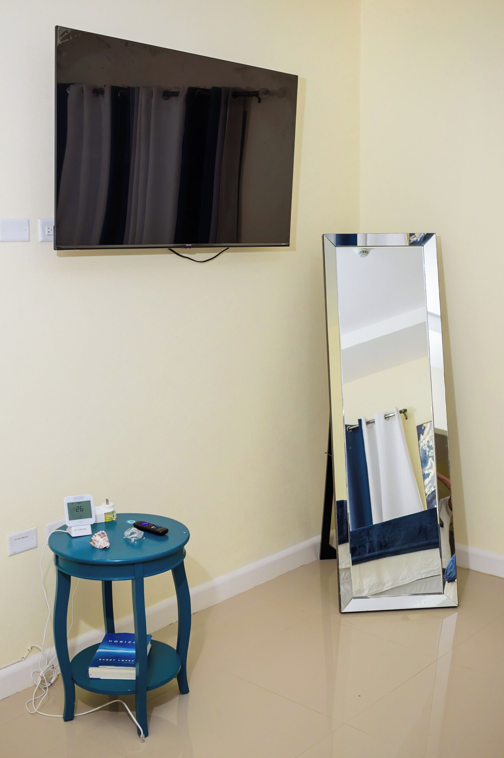 Une pièce dotée d’un téléviseur à écran plat fixé au mur, d’un grand miroir, d’une petite table ronde sur laquelle se trouve un livre, et d’une télécommande.