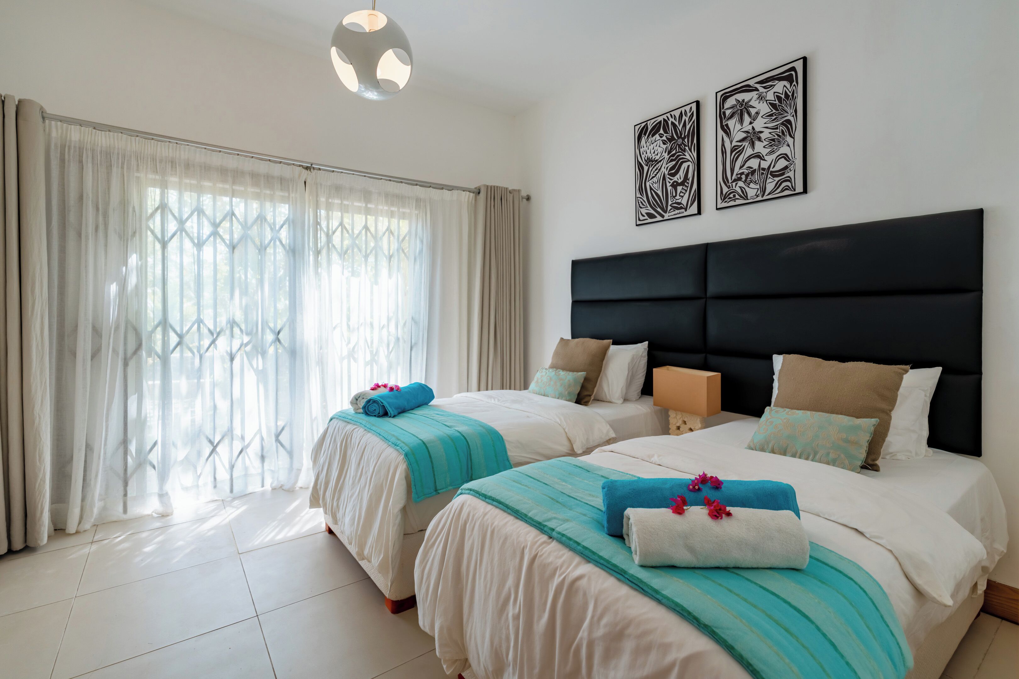 A hotel room with two beds, a large window with curtains, a black headboard, and two wall art pieces.