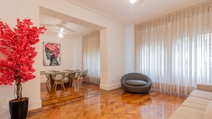 Una sala moderna con piso de madera en diseño herringbone, un sofá gris, un comedor con mesa y sillas, y una planta grande en maceta con flores rojas.