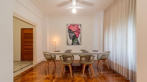 Comedor con una larga mesa dispuesta para seis personas, rodeada de sillas de mimbre, un ventilador de techo y un cuadro en la pared.