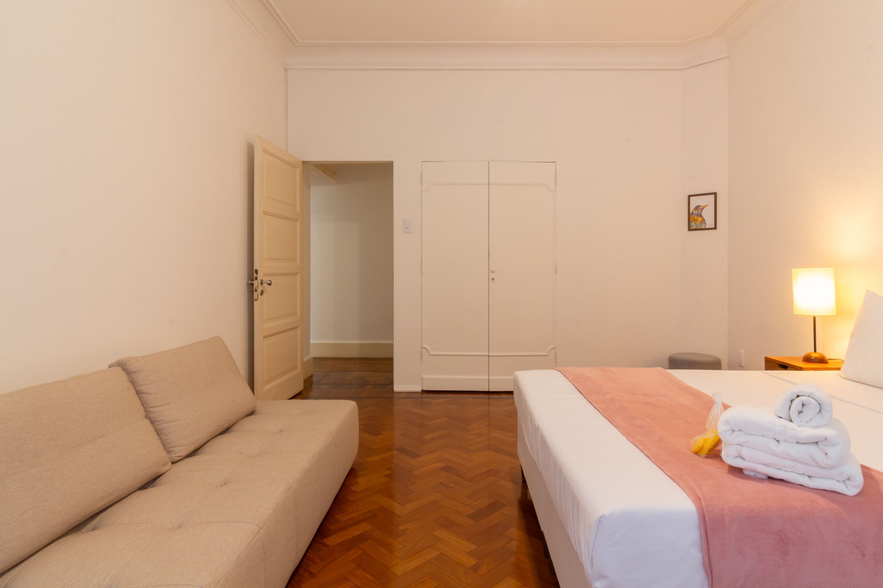 A hotel room with a bed, a sofa, a wardrobe, and a nightstand with a lamp.