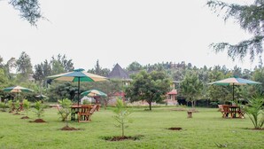 An outdoor seating area with tables and chairs under umbrellas, surrounded by trees and buildings in the background. - Balance Park Hotel  (Kisumu)