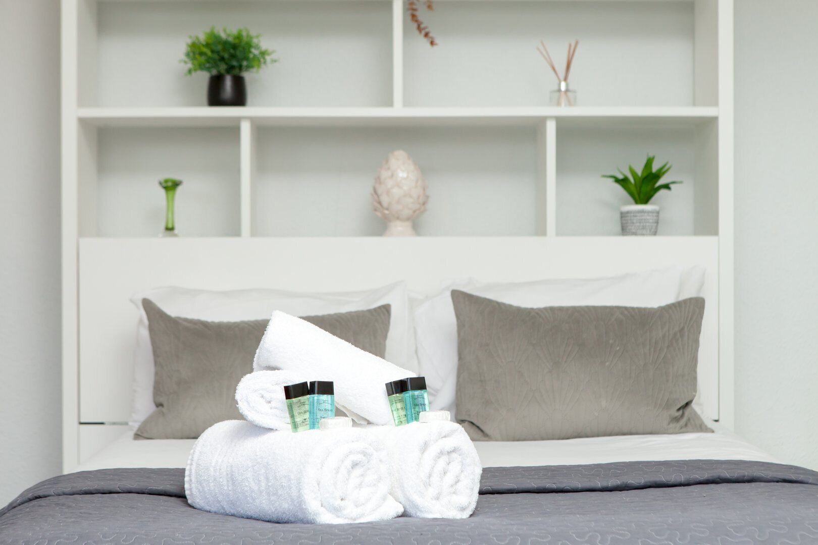 A neatly made bed with white and grey pillows, a grey blanket, and rolled white towels.