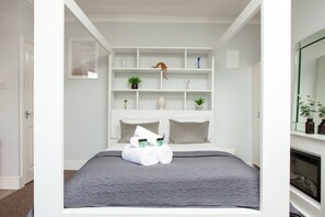 A modern bedroom with a bed, grey bedding, a white headboard, and a built-in shelving unit with decorative items. - Charming Brighton Studio | Calm, Cosy & Central (Brighton)