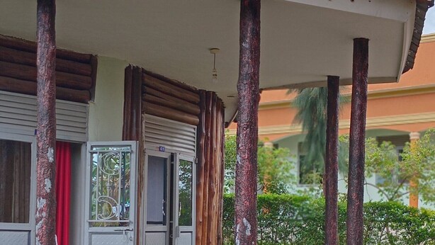 A building with a covered porch supported by wooden pillars, a door with a metal grille, and a red curtain. - rwabs legacy motel mubende (Mubende)