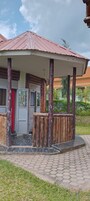Un edificio con un porche cubierto sostenido por pilares de madera, una puerta con celosía metálica y una cortina roja.