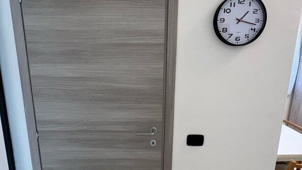 A closed grey door with a keyhole and a lock, a white wall clock, and a small black switch. - LITTLE HOUSE (Fossarmato)