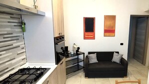 A compact living space with a kitchenette, a black sofa, and two abstract paintings on the wall. - LITTLE HOUSE (Fossarmato)
